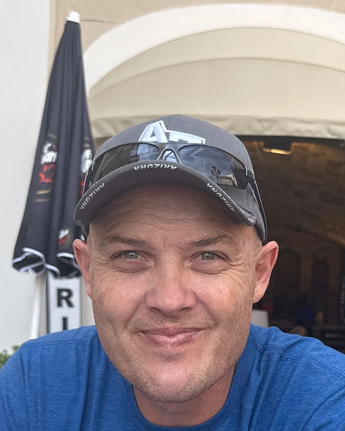Maximus Sego, seated outside a café in Italy, wearing a cap with sunglasses pushed up on it, looking at the camera.